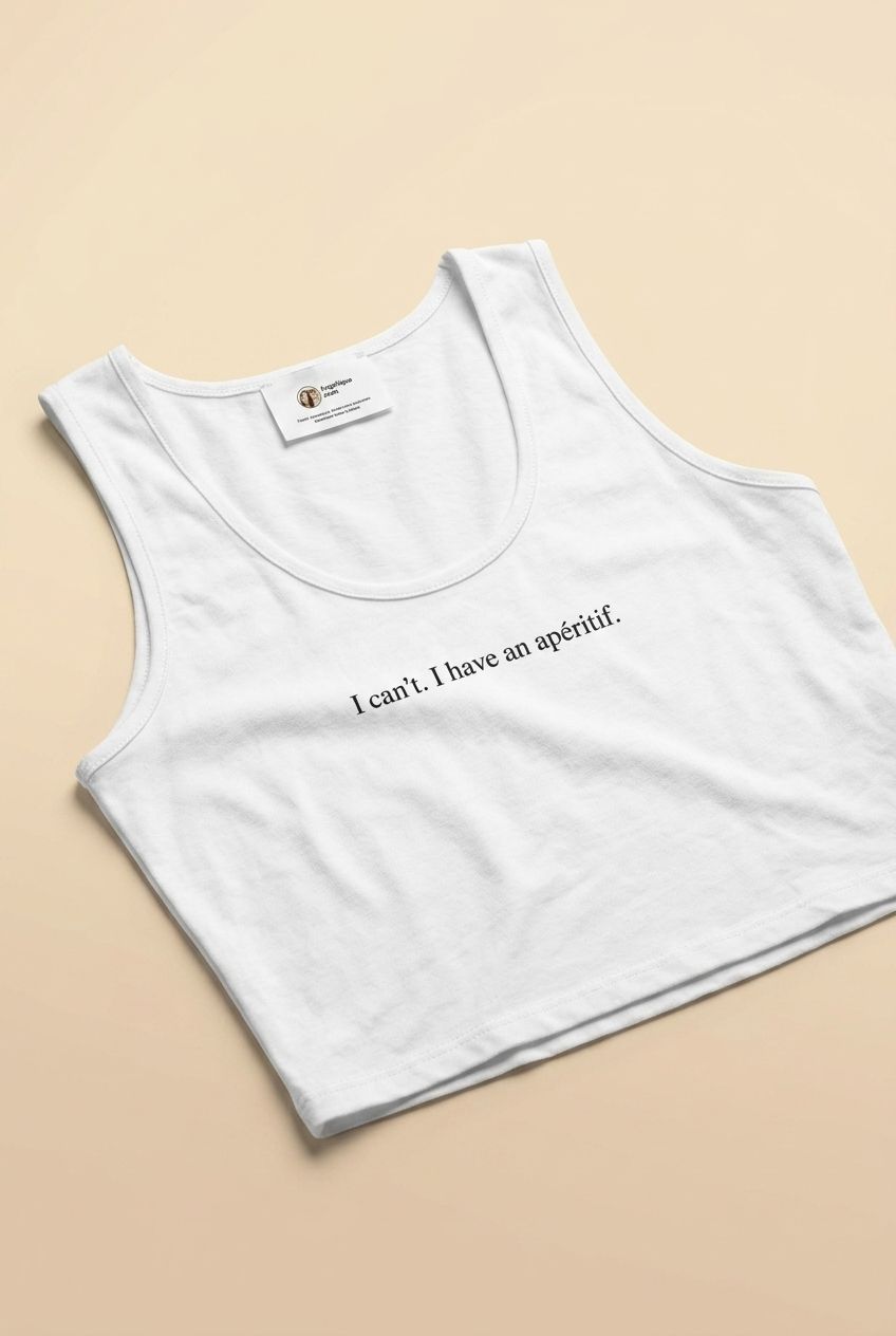 Model wearing a white 'I can't. I have an apéritif.' crop top, shown on a neutral background with soft natural lighting, styled casually.