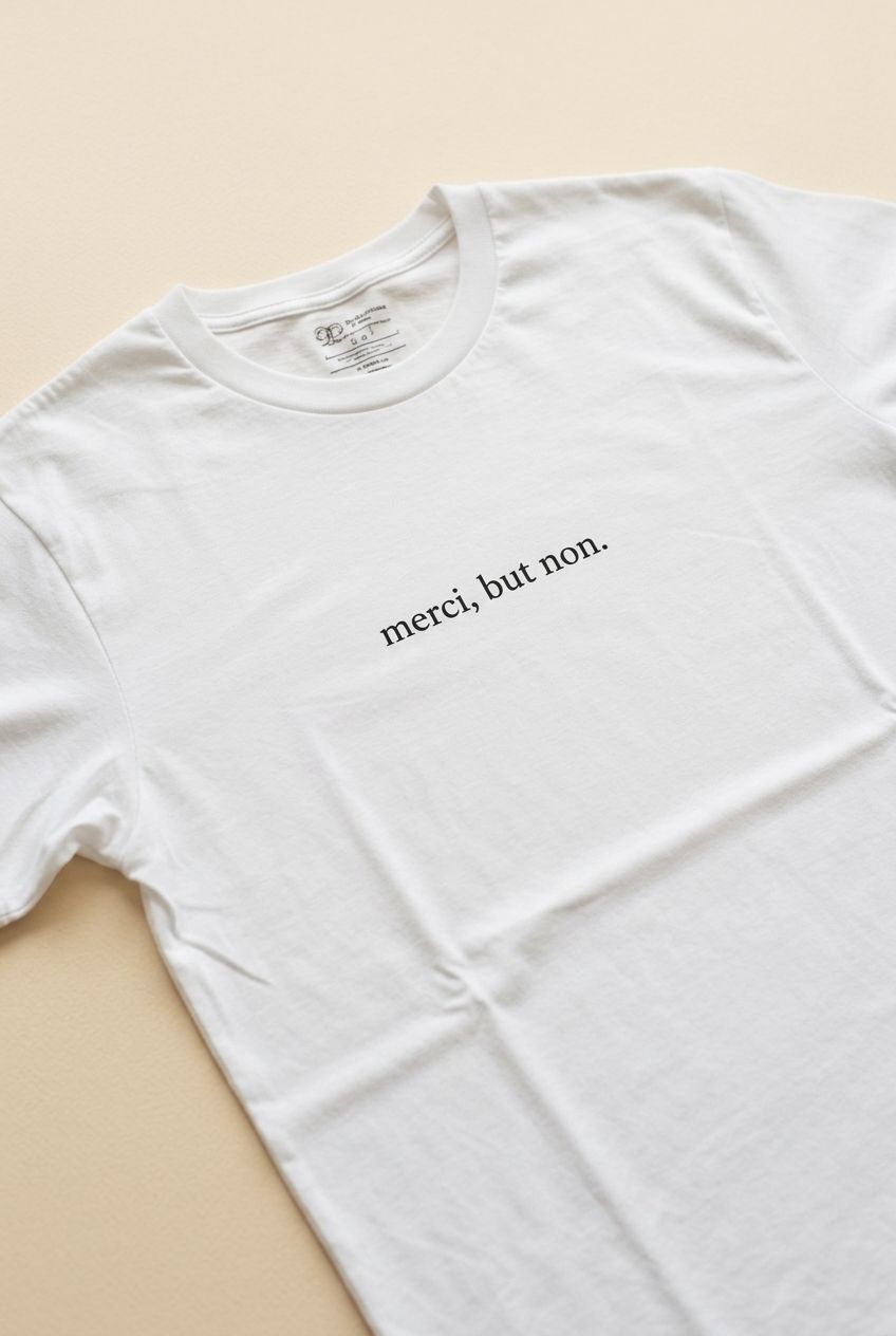 Light unisex T-shirt with 'merci, but non' quote, centered on a natural drape in a soft white studio setting.