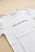Front view of white cotton unisex t-shirt with centered graphic text, placed on a wooden table in soft natural light.